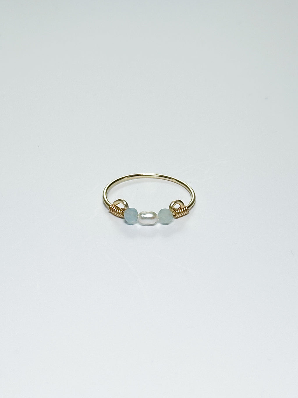 Gold wrapped Ring with Pearl in the middle and two light blue beads on two sides on a white background with shadows.