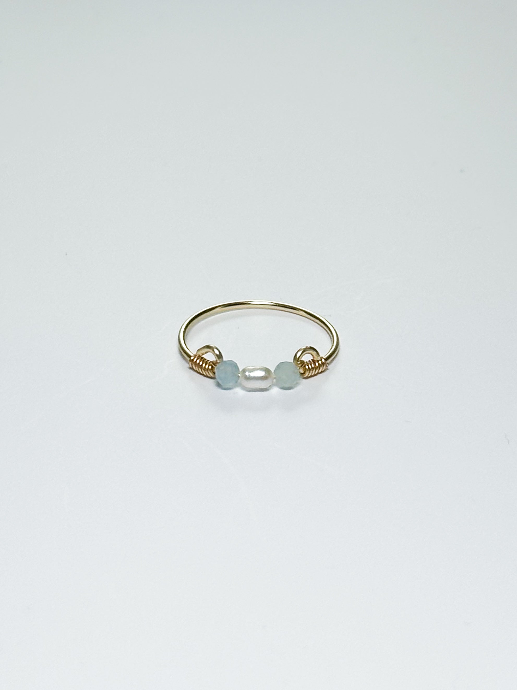 Gold wrapped Ring with Pearl in the middle and two light blue beads on two sides on a white background with shadows.
