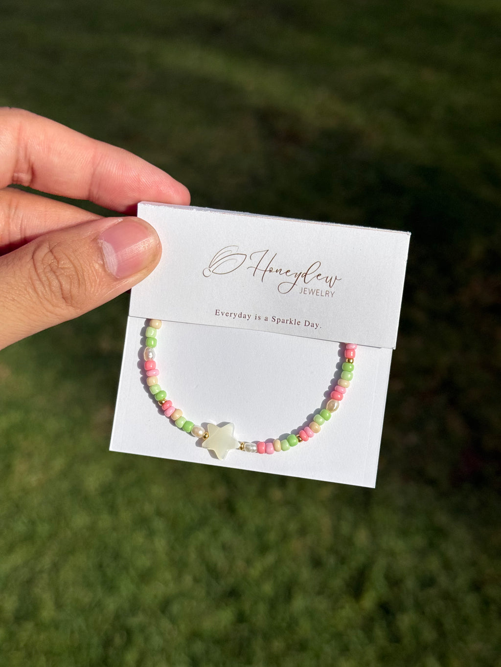 Hand holding a star-shaped mother of pearl bracelet colourful (pink, green and beige) beads on a card with 'Honeydew' branding against a grassy background.