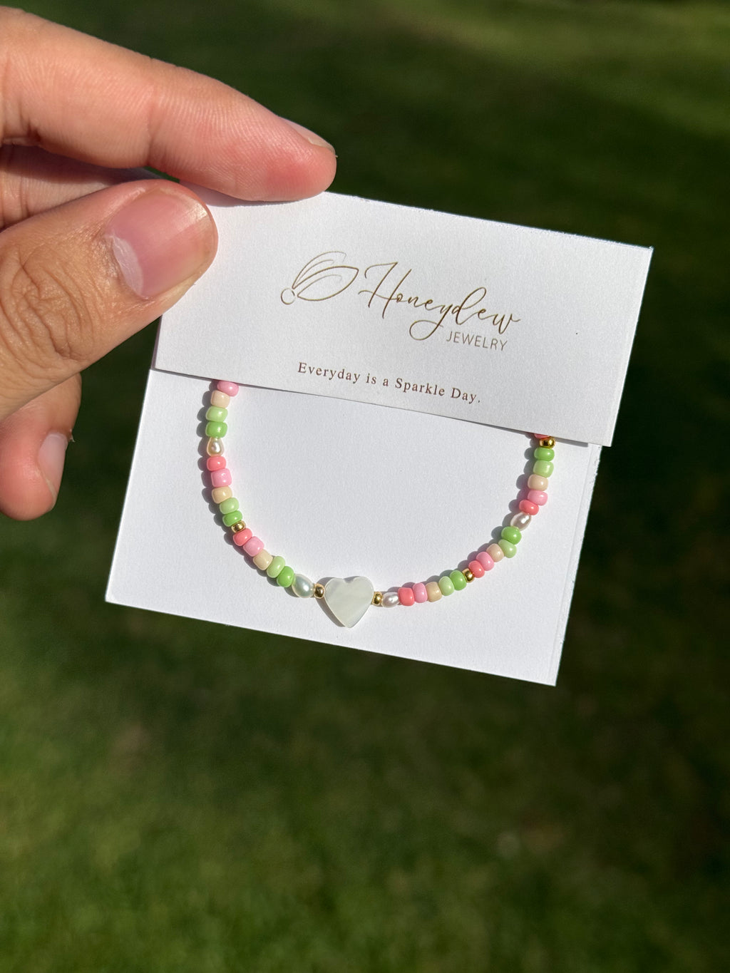 Hand holding a bracelet with heart-shaped mother of pearl and colourful (green, beige, and pink) beads, on a Honeydew Jewellery card against a grassy background.