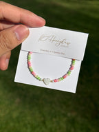 Hand holding a bracelet with heart-shaped mother of pearl and colourful (green, beige, and pink) beads, on a Honeydew Jewellery card against a grassy background.