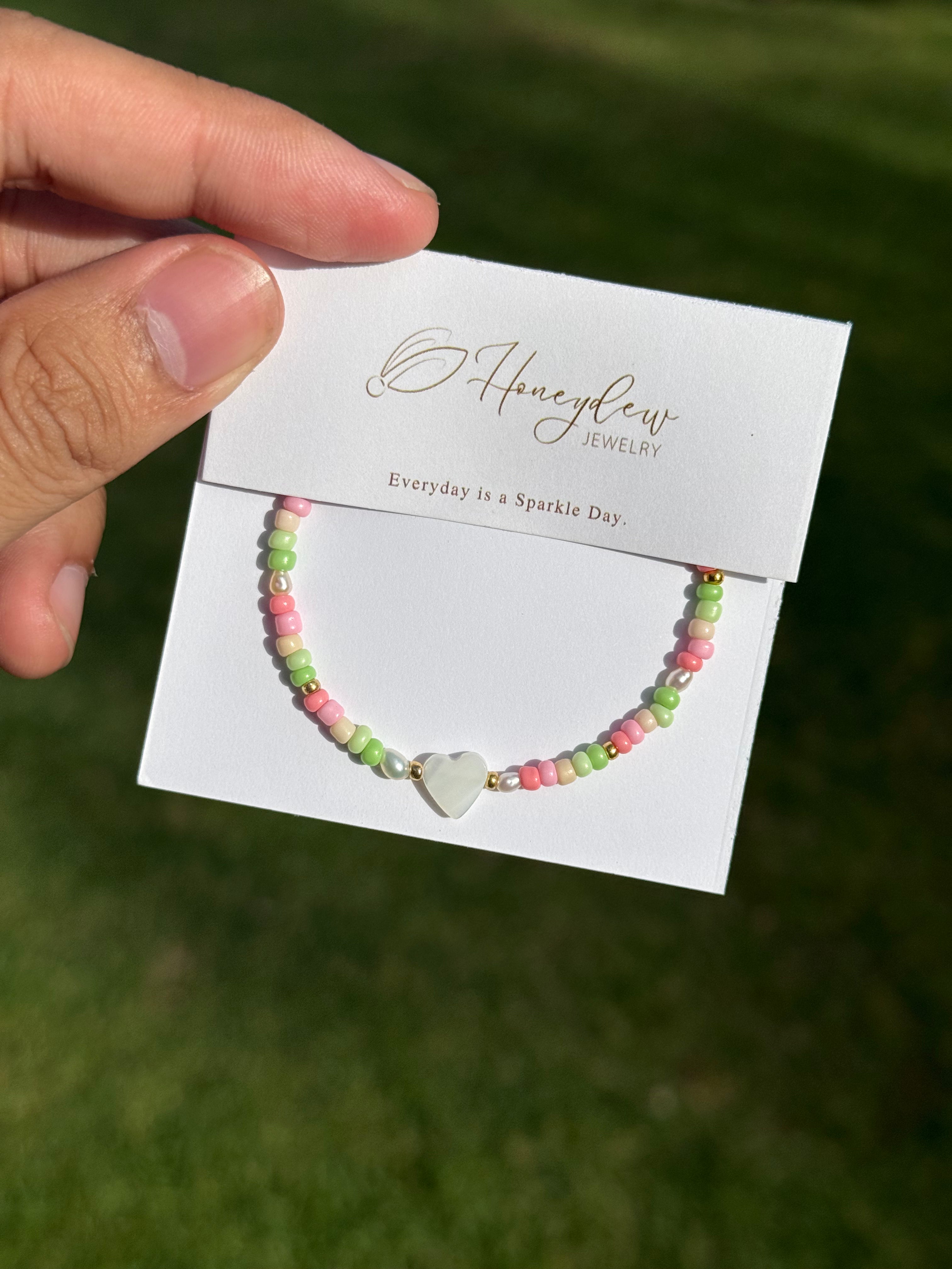 Hand holding a bracelet with heart-shaped mother of pearl and colourful (green, beige, and pink) beads, on a Honeydew Jewellery card against a grassy background.
