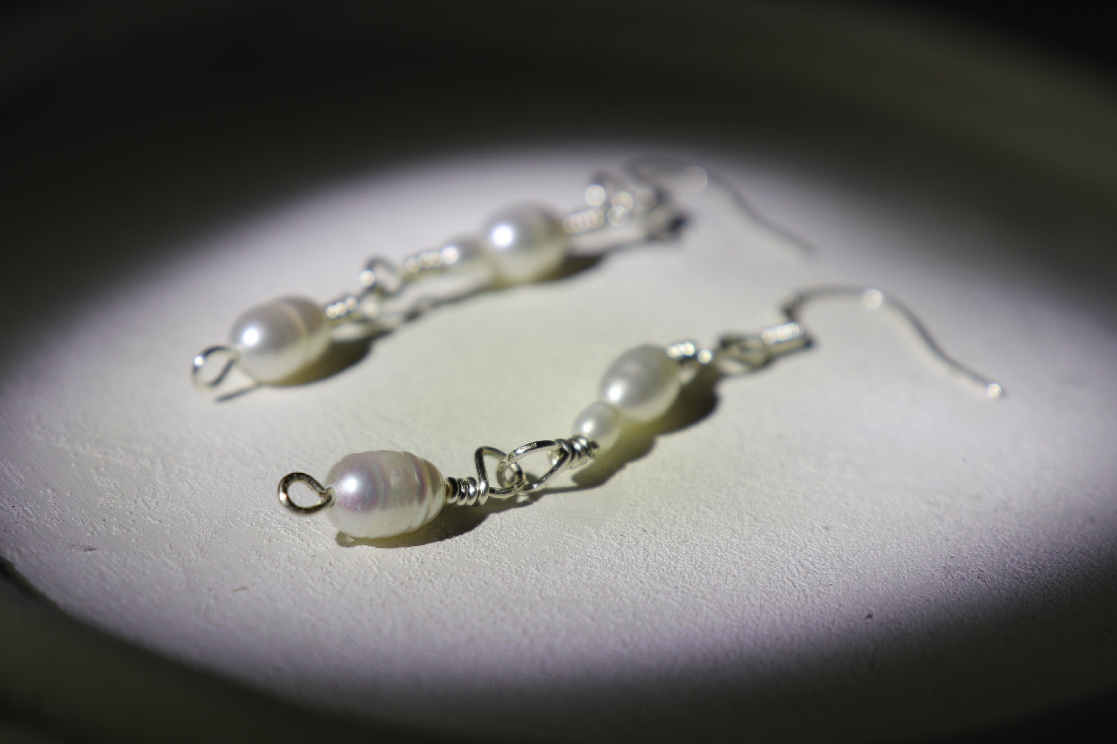 Pearl link silver earrings on a dark background with a spotlight effect, with zoomed effects detailing the hooks and pearls of one of the earrings