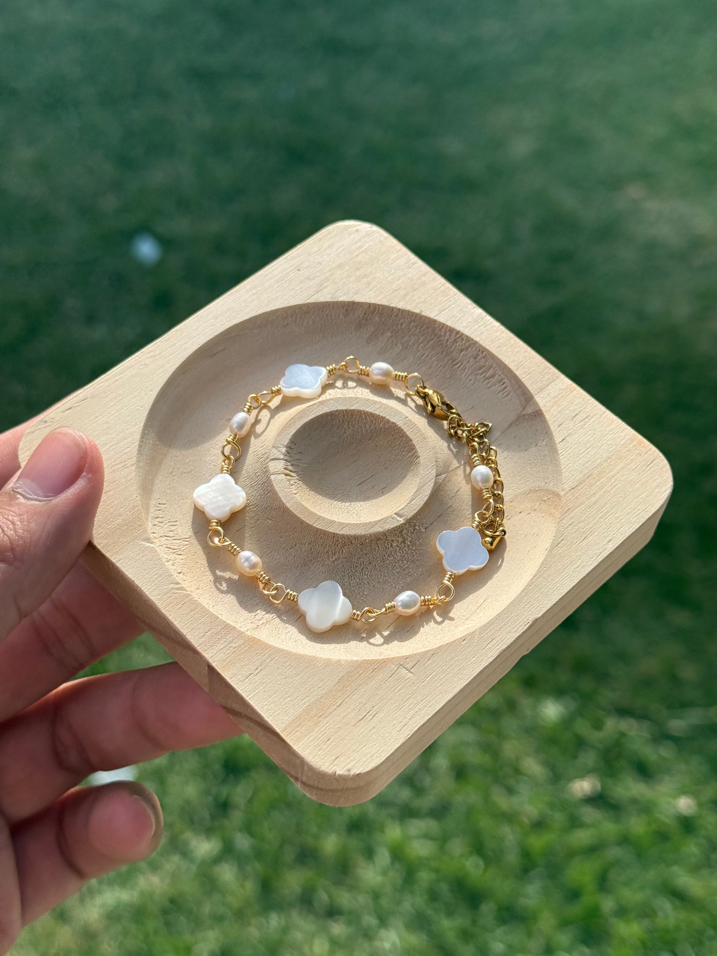 Gold Clover Pearl and Mother of Pearl Bracelet on a display tray held by a hand against a green grass outdoor background.