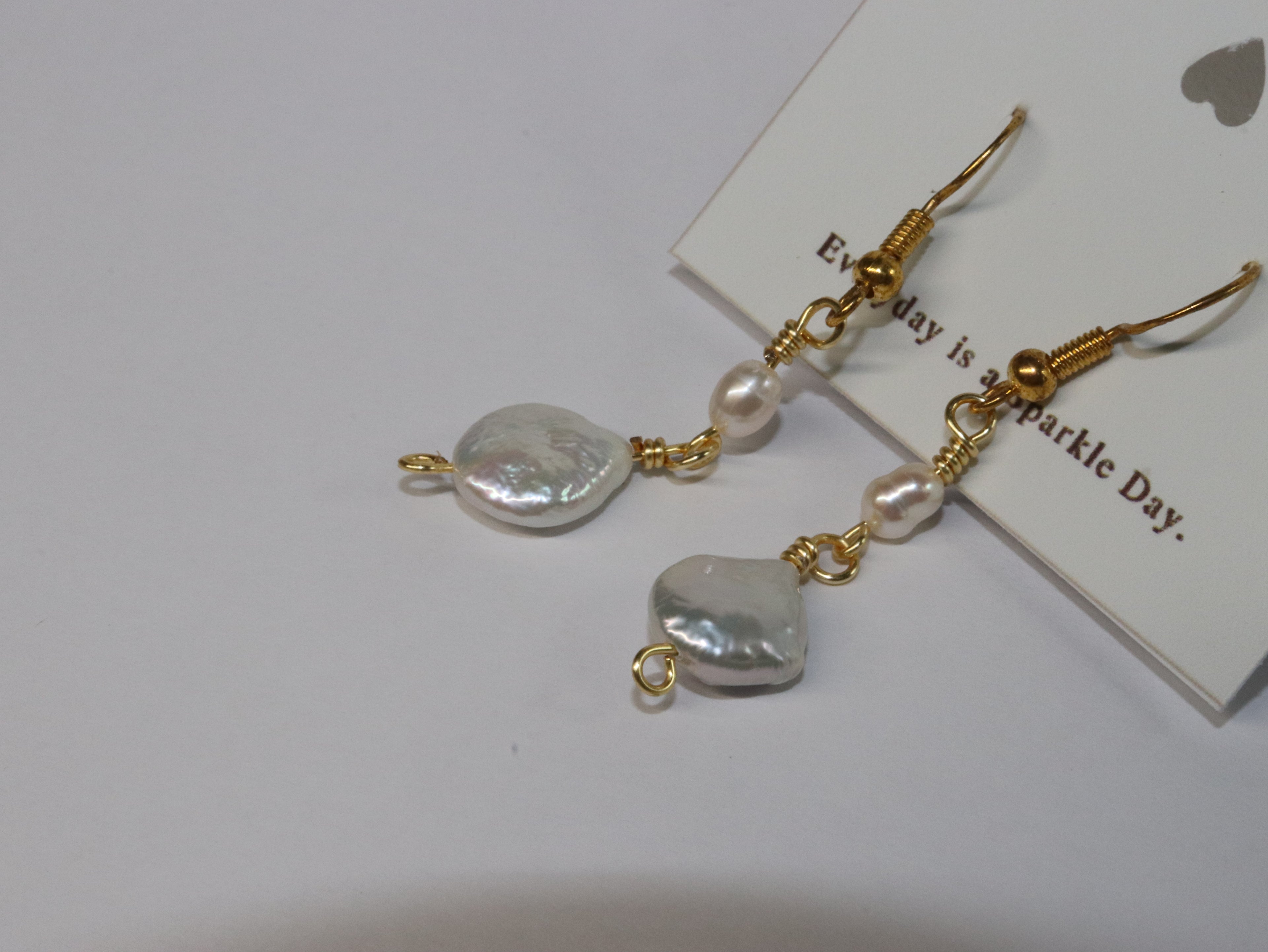 A close up shot of Gold link earrings with pill-shaped pearl and round, plate-shaped pearl, on a white card with Honeydew's slogan "Everyday is a Sparkle Day" with white background.