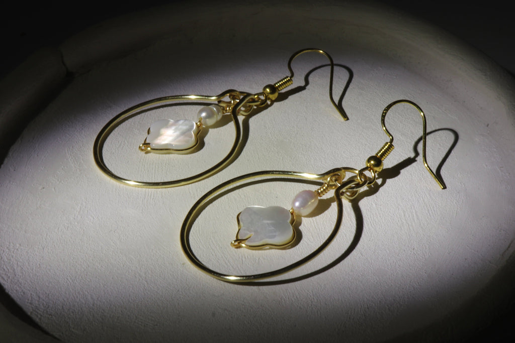 Gold hoop earrings with clover-shaped mother of pearl and freshwater pearl placed on a white clay plate, on a dark background.