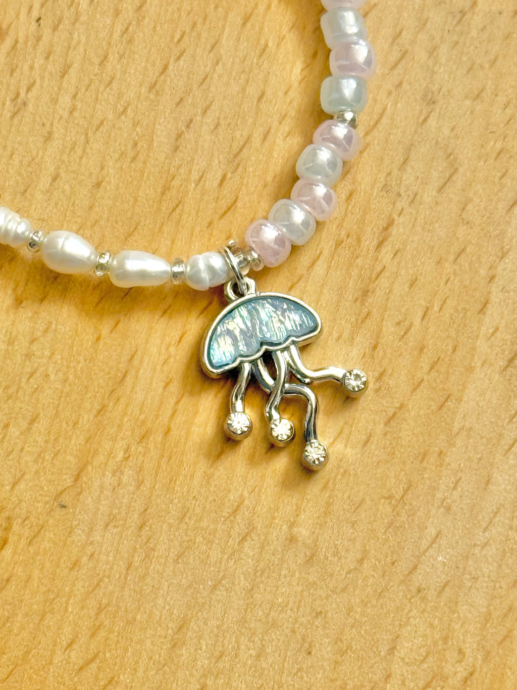 A close up half shot of a half-pearl half-beads (blue, pink, white) bracelet with the focus on the jellyfish charm, on a wooden coaster.