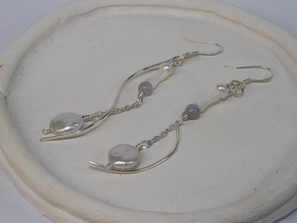 Silver-plated pair of earrings with silver-plated hooks on a white ceramic plate. The body consists of a pearl, a cone-shaped mother of pearl, and a purple bead, connected by a chain and round, plate-shape pearls at the tail. A separate curvy metal body also follow along the main body.