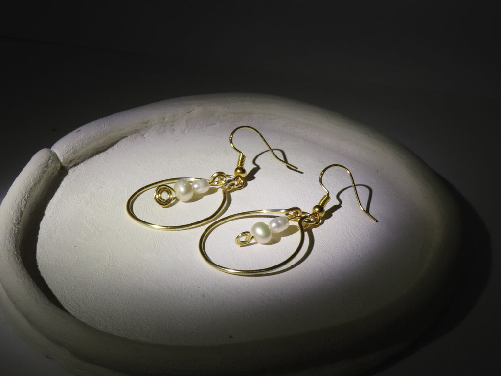 Gold hoop pearl earrings with two pearls on an off-white ceramic plate, with dark background a spotlight shining on the earrings.