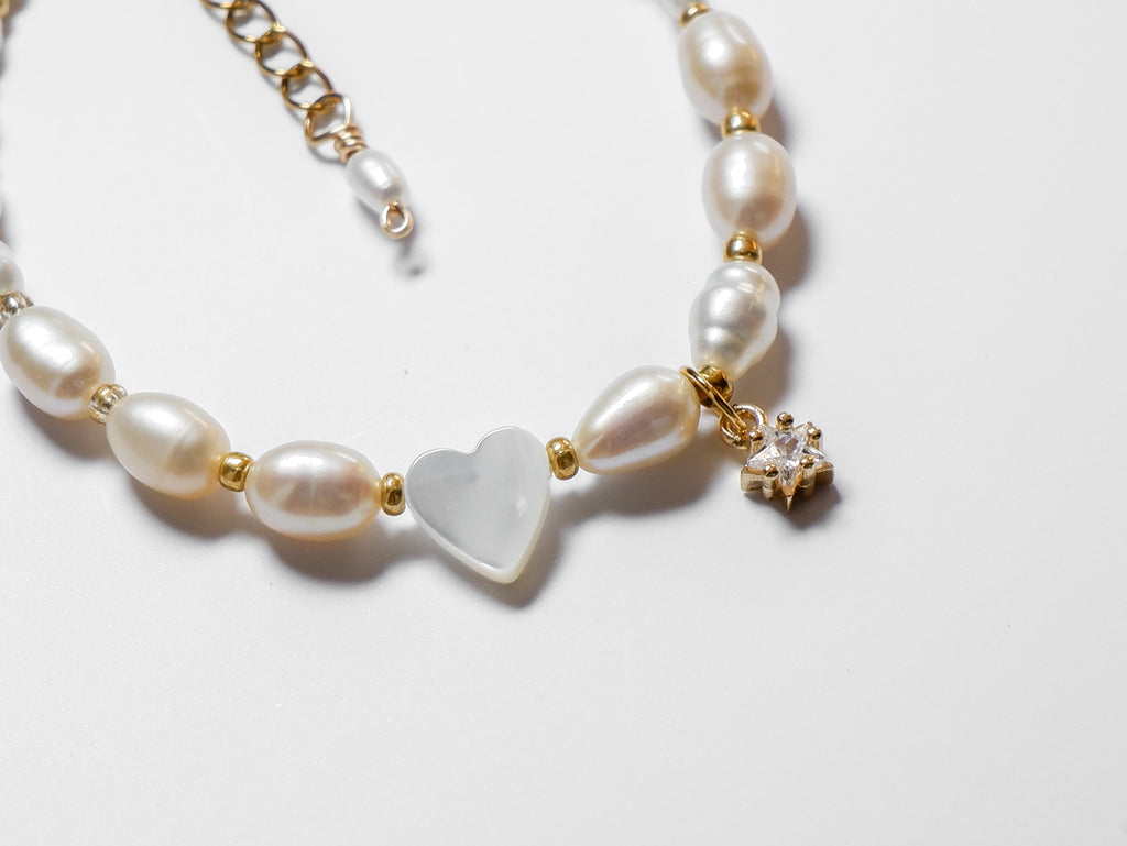 An ultra close-up half-shot of a gold pearl bracelet, with an off-centre heart-shaped mother of pearl and gold star charm lying on a white background.