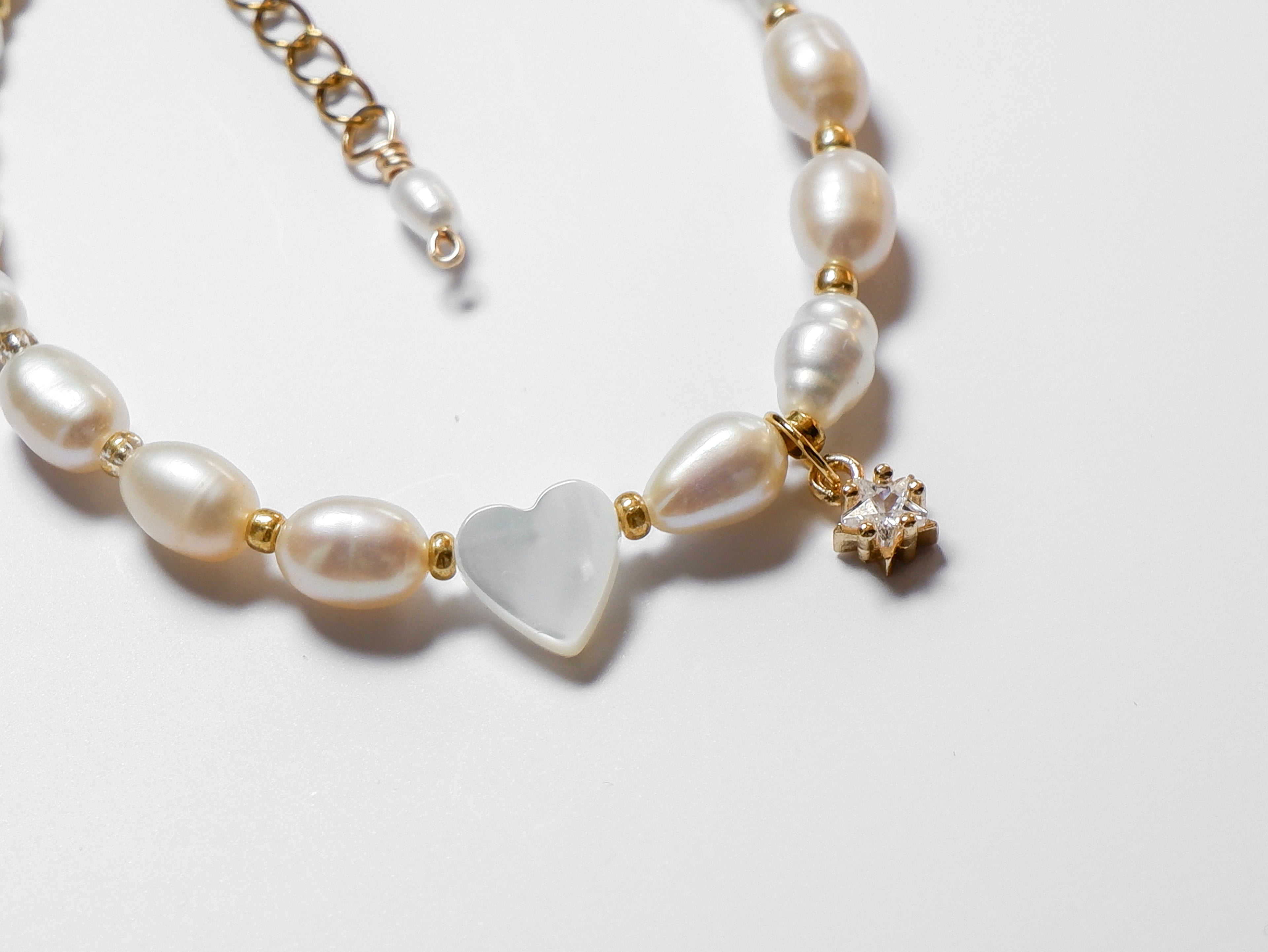An ultra close-up half-shot of a gold pearl bracelet, with an off-centre heart-shaped mother of pearl and gold star charm lying on a white background.