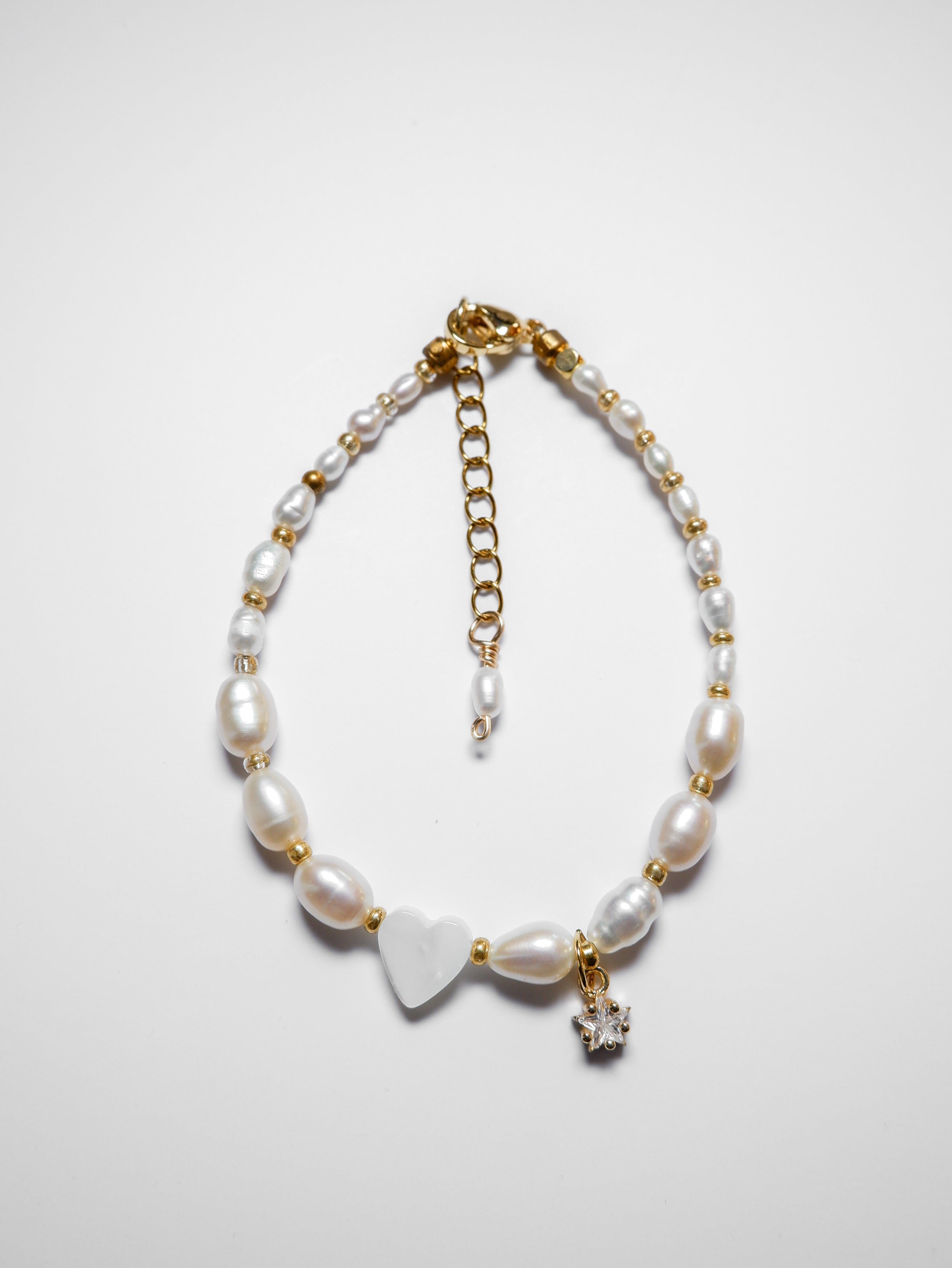 A gold pearl bracelet, with an off-centre heart-shaped mother of pearl and gold star charm on a white background.
