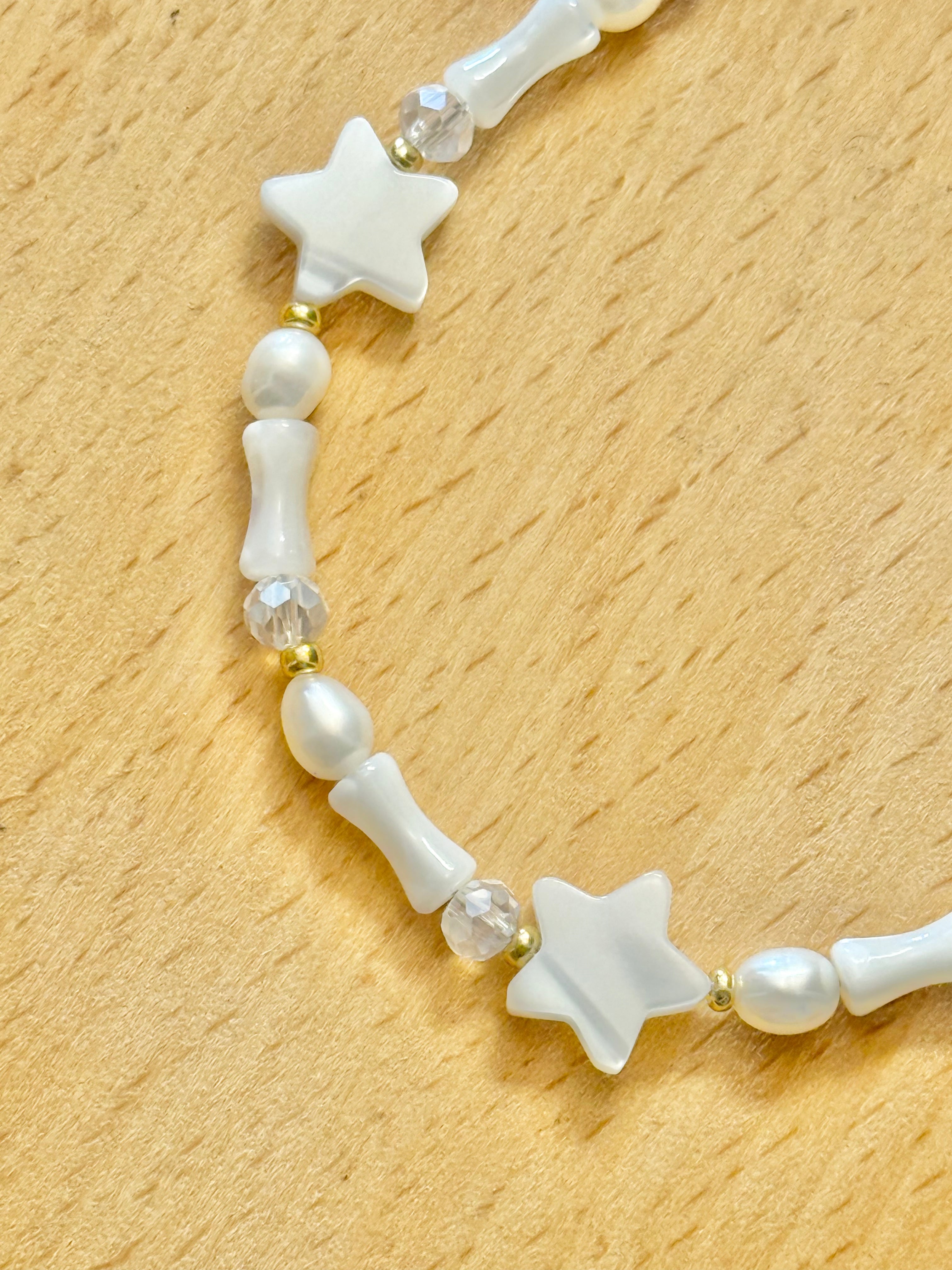 A close up half shot of a bracelet with star-shaped and cone-shaped mother of pearls, round pearls and clear beads, on a wooden coaster.
