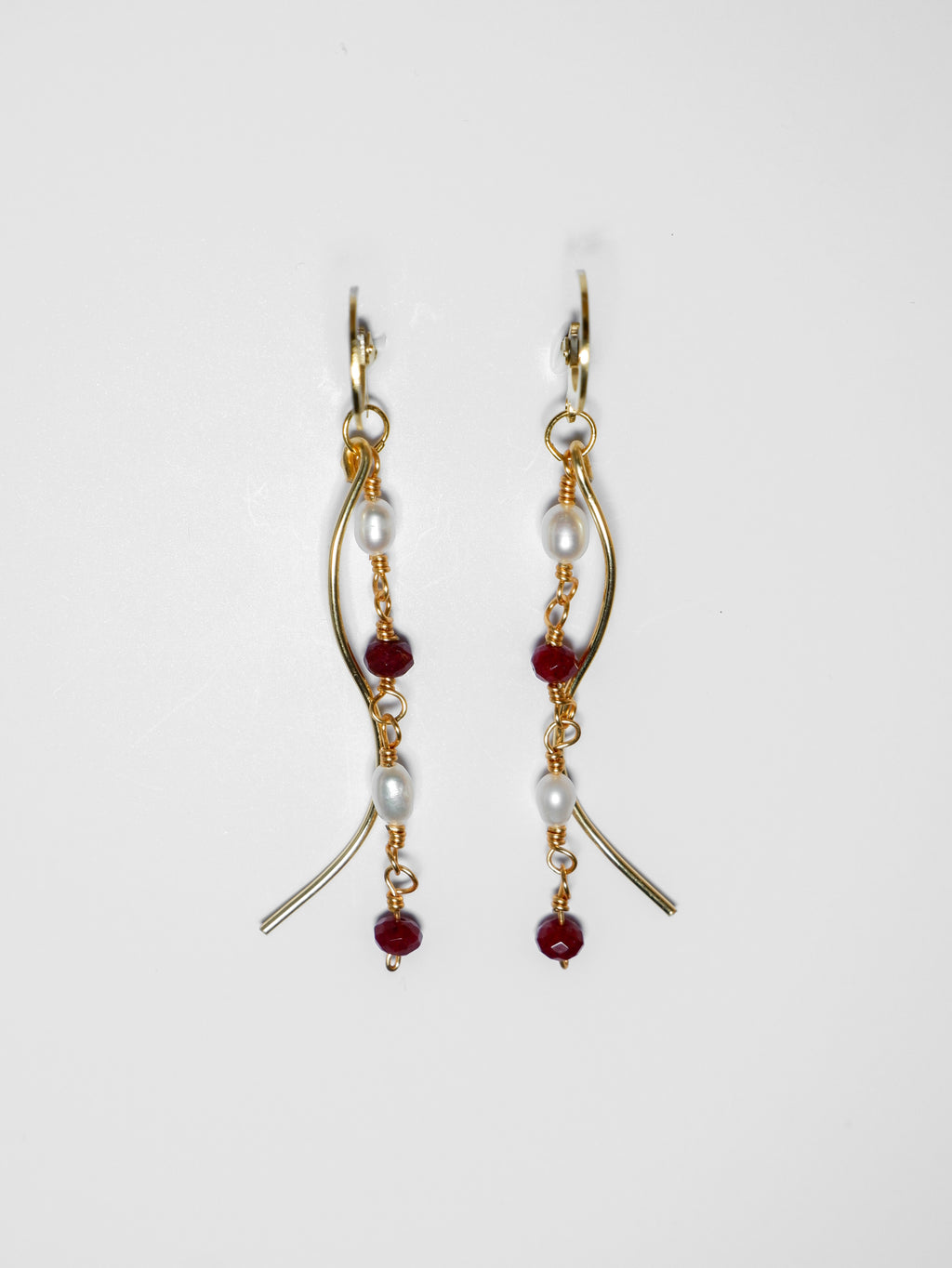 Gold pearl earrings with two bodies–one with crimson red beads and pearls linked by hooks, another is a curvy gold-plated body, on a white background.