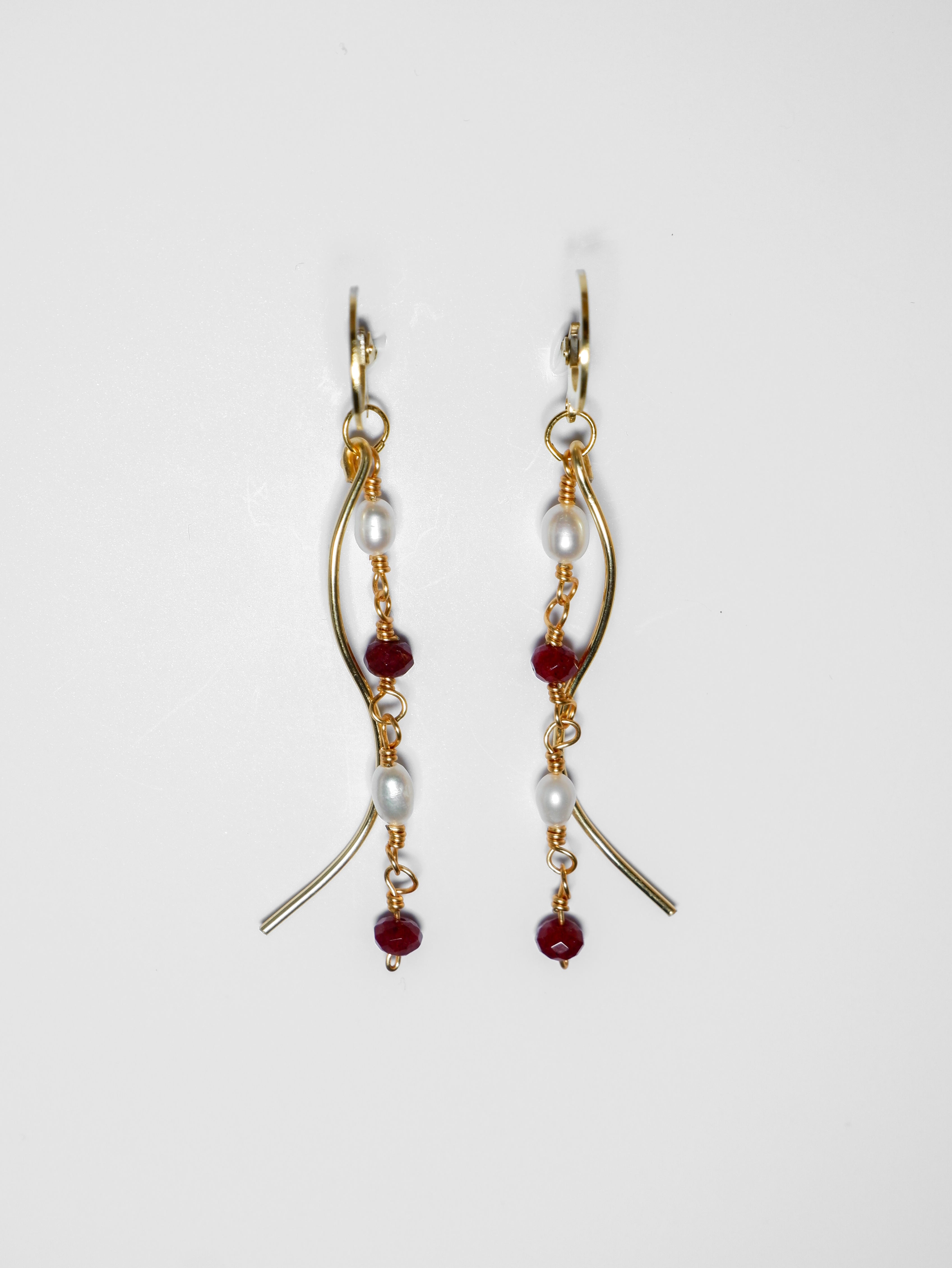 Gold pearl earrings with two bodies–one with crimson red beads and pearls linked by hooks, another is a curvy gold-plated body, on a white background.