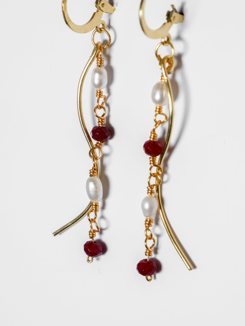 An ultra close-up shot of a Gold pearl earrings with two bodies–one with crimson red beads and pearls linked by hooks, another is a curvy gold-plated body, on a white background.