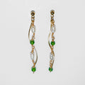 Gold pearl earrings with two bodies–one with emerald green beads and pearls linked by hooks, another is a curvy gold-plated body, on a white background and studio backdrop.