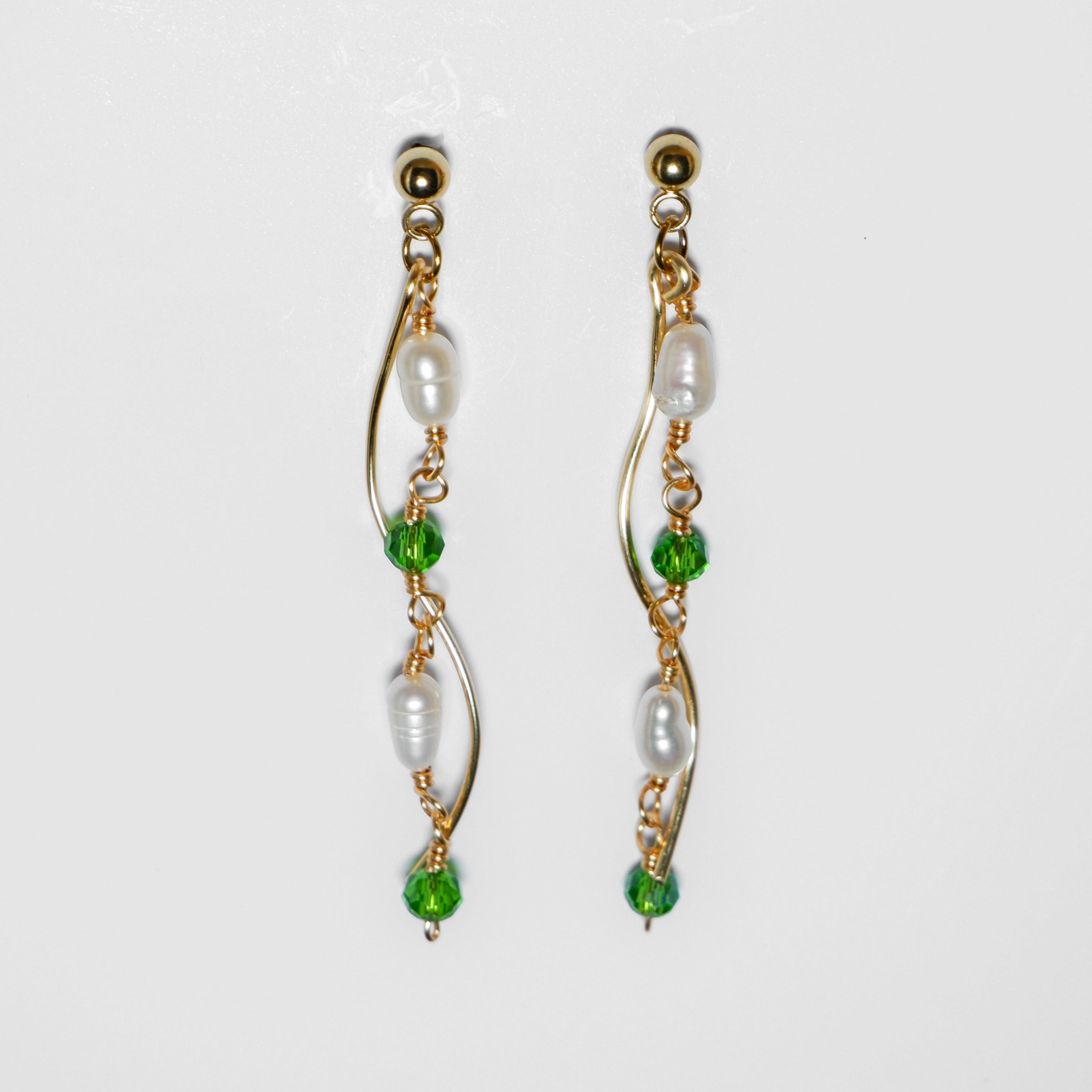 Gold pearl earrings with two bodies–one with emerald green beads and pearls linked by hooks, another is a curvy gold-plated body, on a white background and studio backdrop.